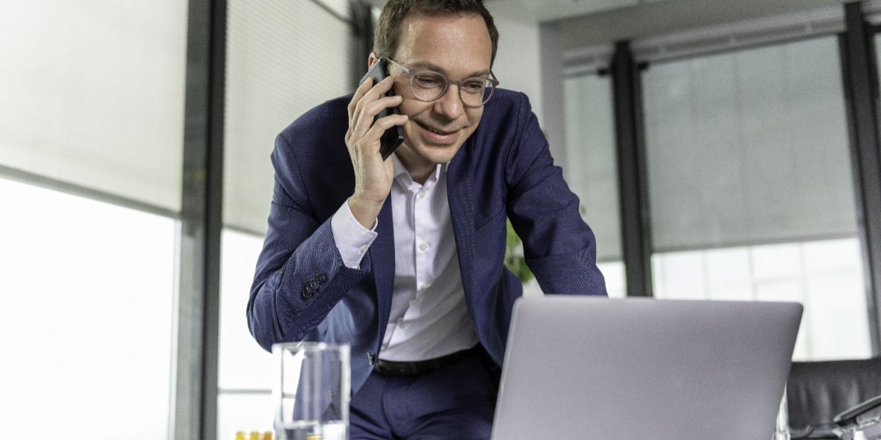 Manager standing behind office desk with laptop computer, holding mobile phone, CRM